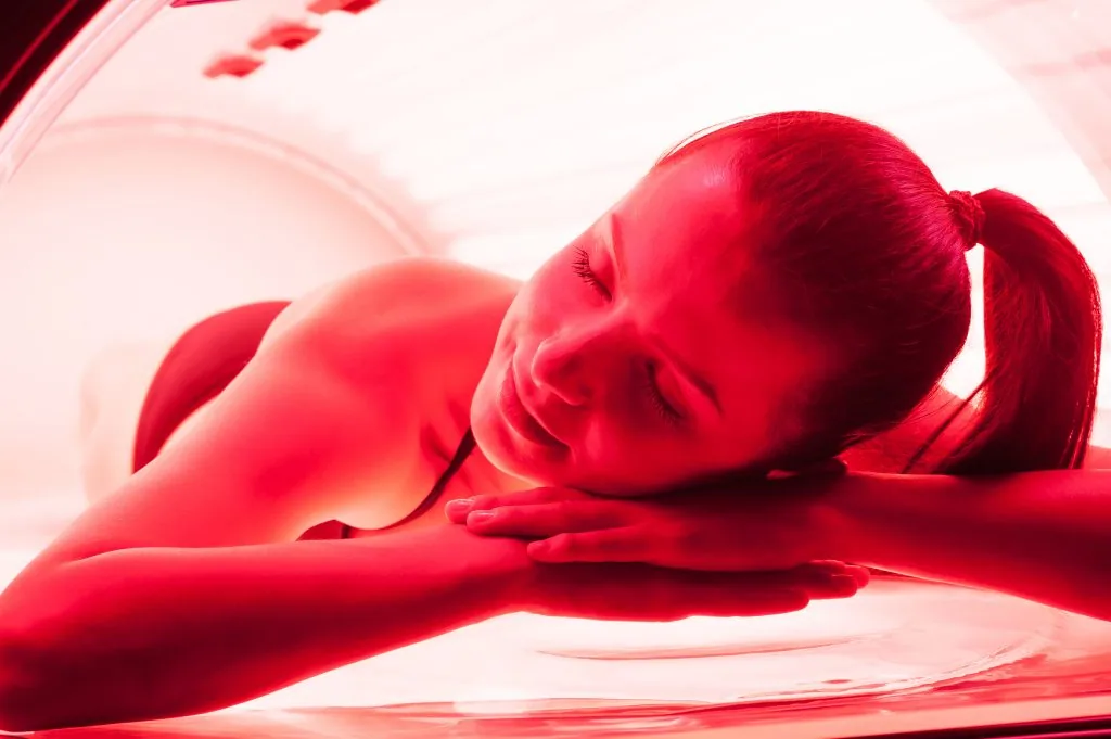 A person lying face down inside a whole body red light bed, skin lit by the red LED panels